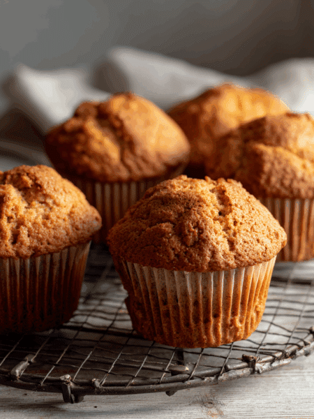 Freshly baked spelt breakfast muffins, batch of six, golden tops, soft crumb texture, placed on a wire cooling rack on a light wooden table.