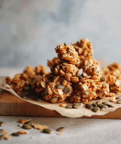 Golden spelt granola clusters arranged on a parchment-lined board. Crisp, irregular clusters with visible nuts, spelt flakes, and pumpkin seeds.