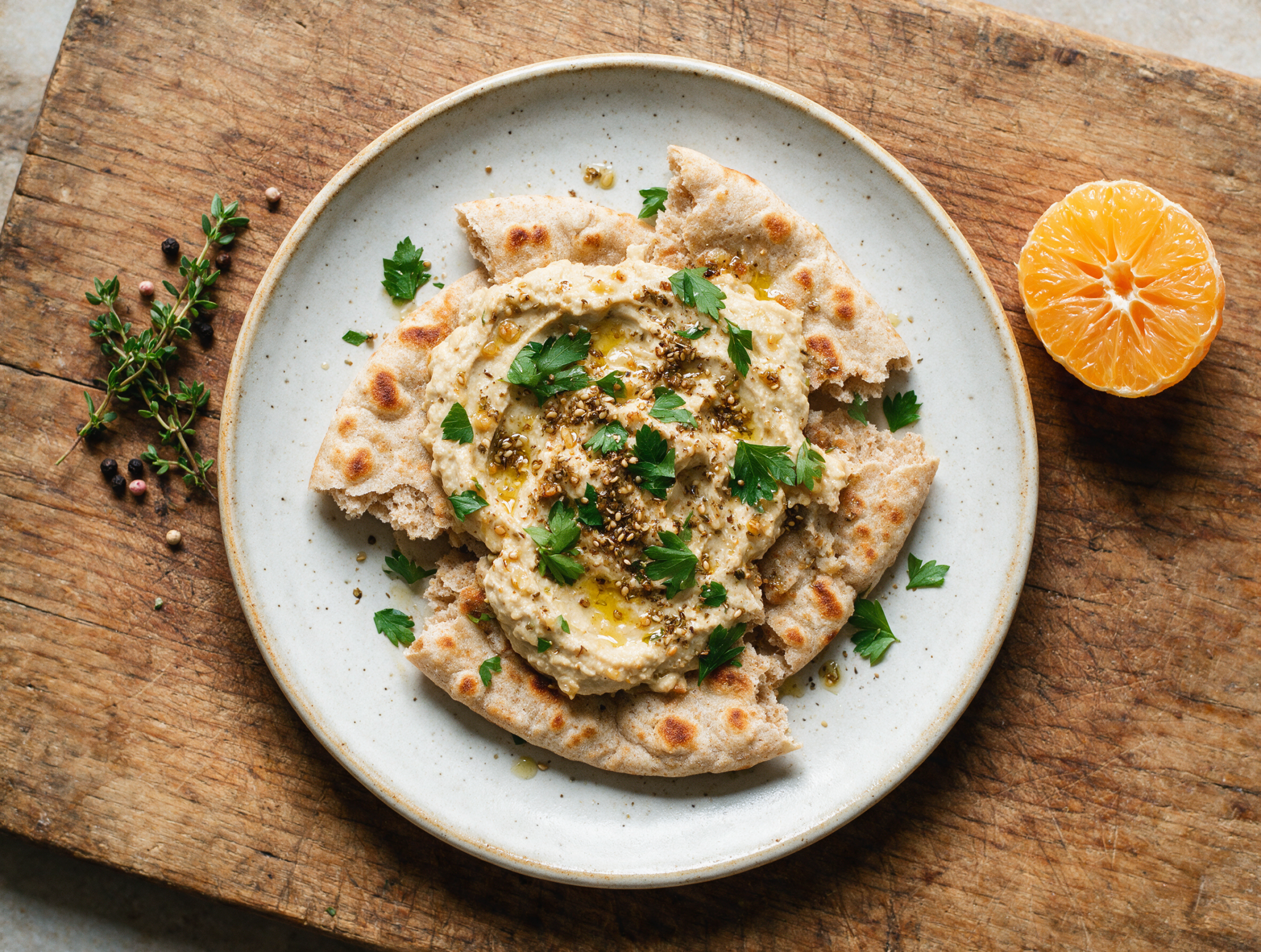 Overhead shot of a torn spelt flatbread spread with roasted garlic hummus, olive oil pooling, za'atar scattered on a wooden board.