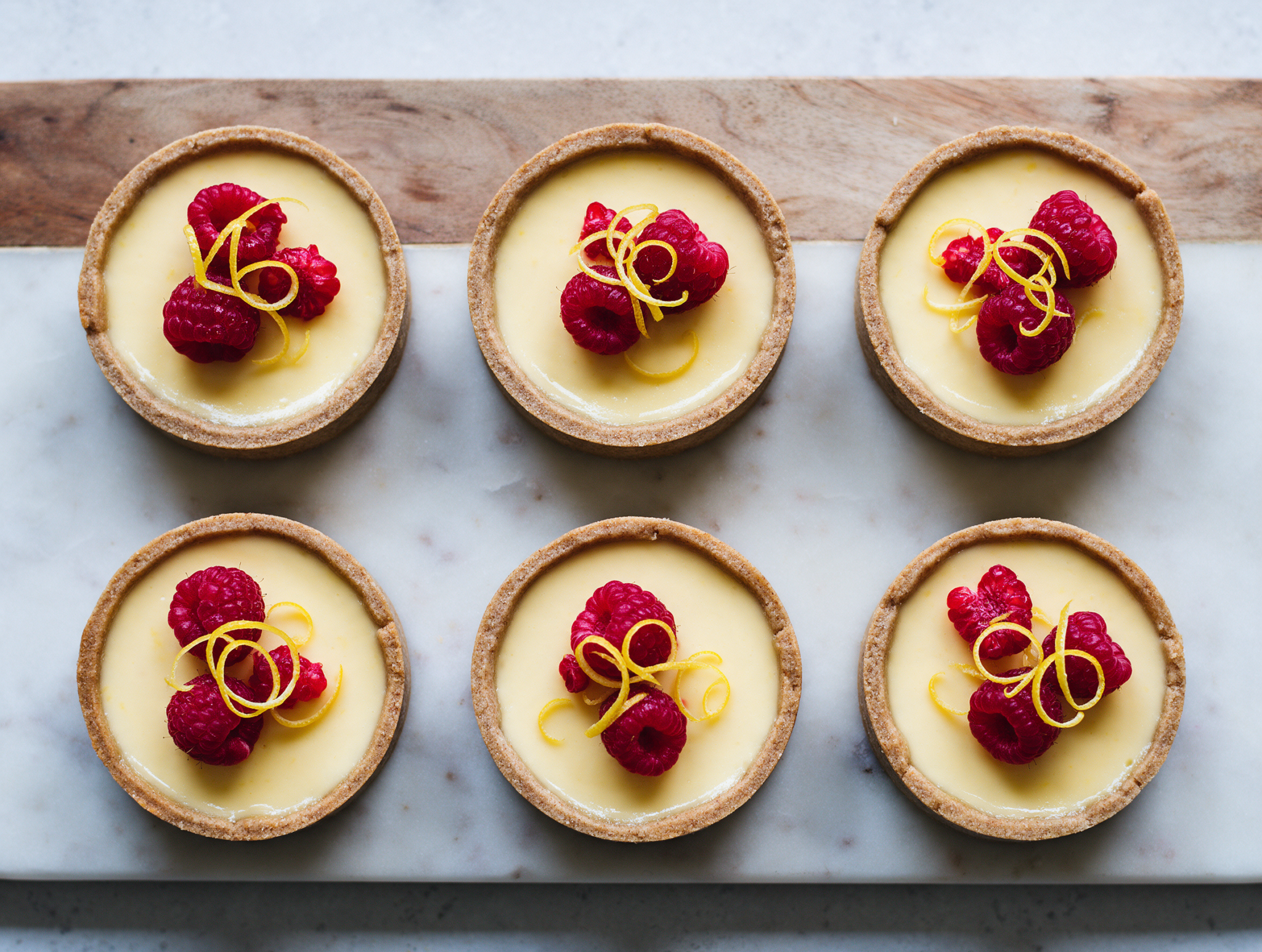 Six golden spelt tartlets with pale lemon posset filling and fresh raspberries arranged on a marble board