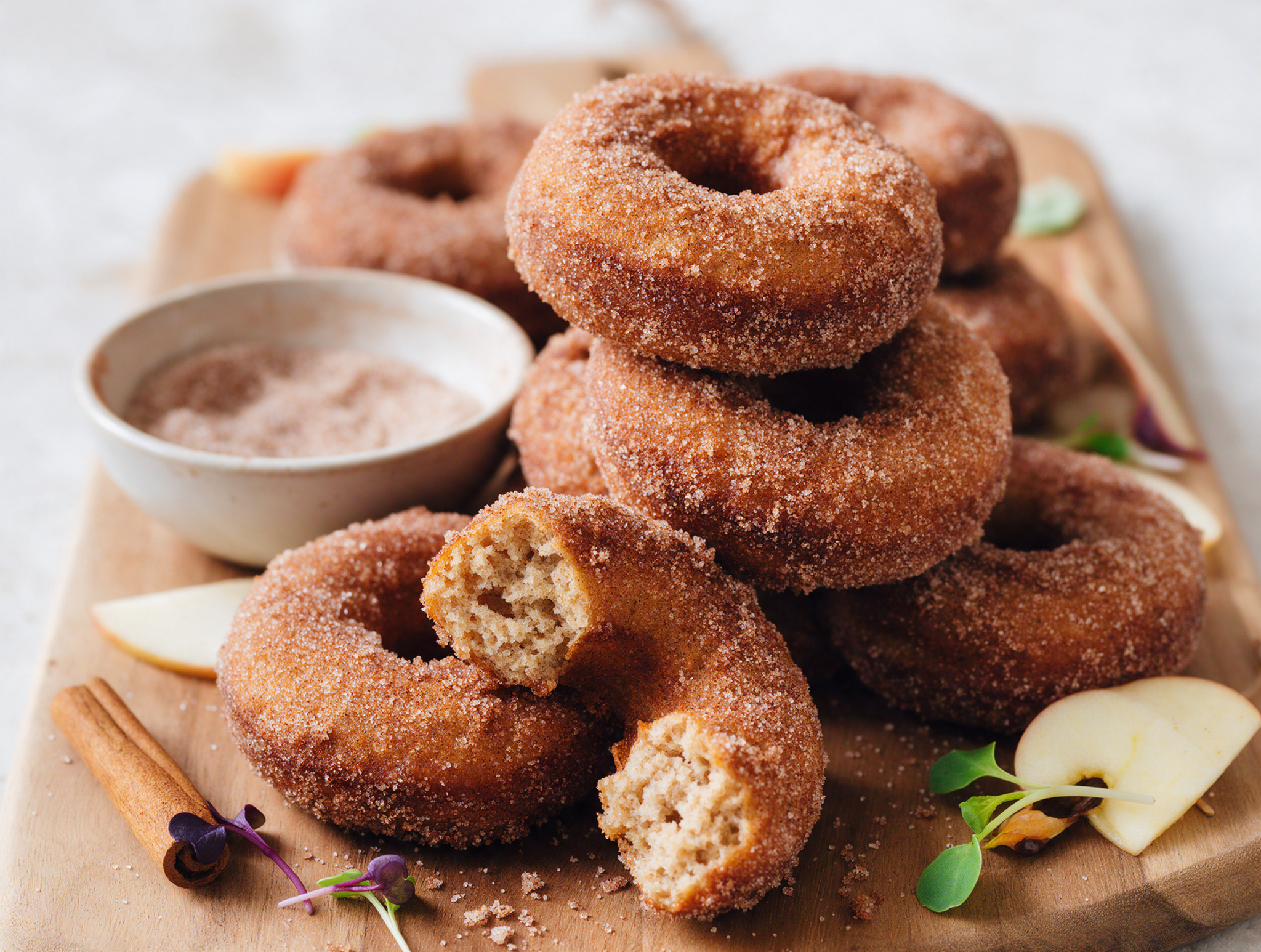 Stack of spelt apple cider pancake doughnuts coated in cinnamon sugar on a wooden board with one split open