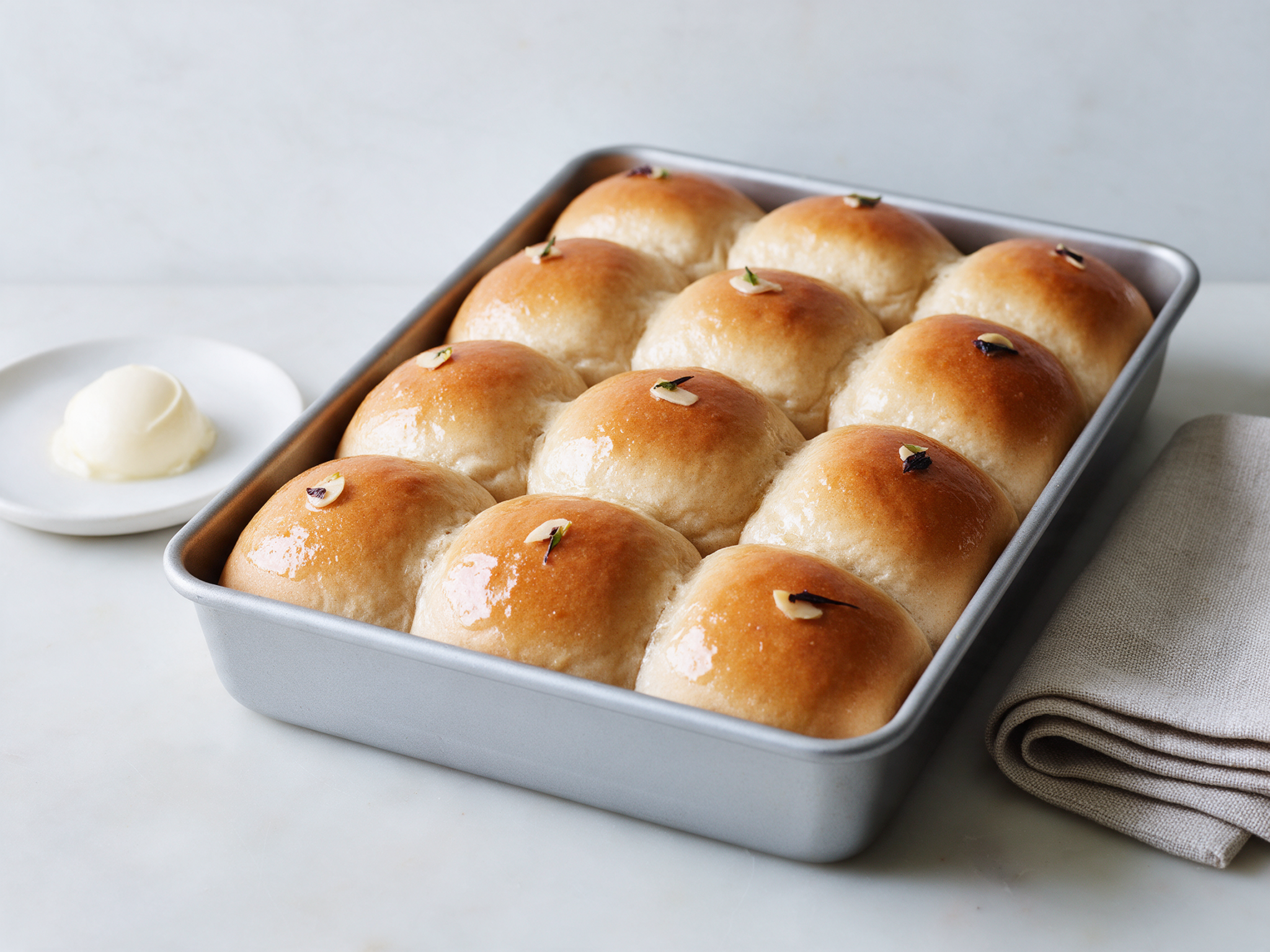 Twelve golden spelt milk buns in a baking pan, glossy with butter, on a linen cloth in window light